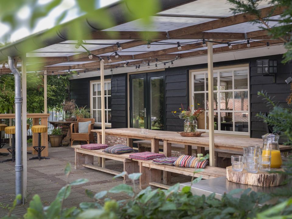 Overdekt terras met tafels en zitbanken met kleurrijke kussens en wat beplanting.