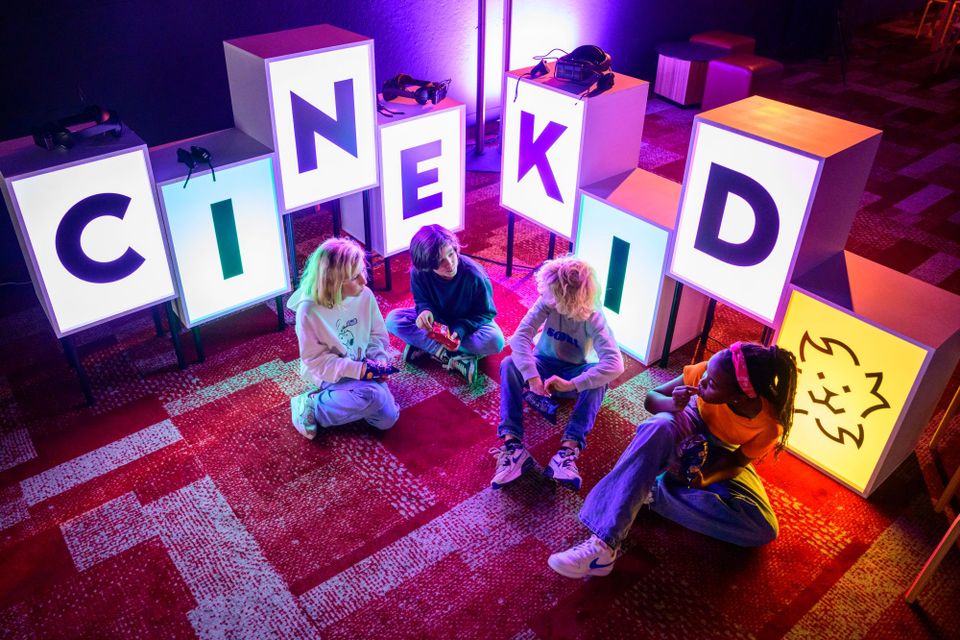 4 kinderen zitten op de grond en kletsen met elkaar. Om hen heel staan lichtgevende letterboxen met de letters C-I-N-E-K-I-D.