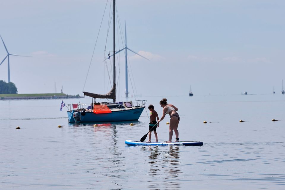 Een jongen en een meisje staan op een supboard op het IJsselmeer bij het strand van Lemmer. Er ligt een lijn met boeien in het water en op de achtergrond ligt een zeilboot en staan windmolens.