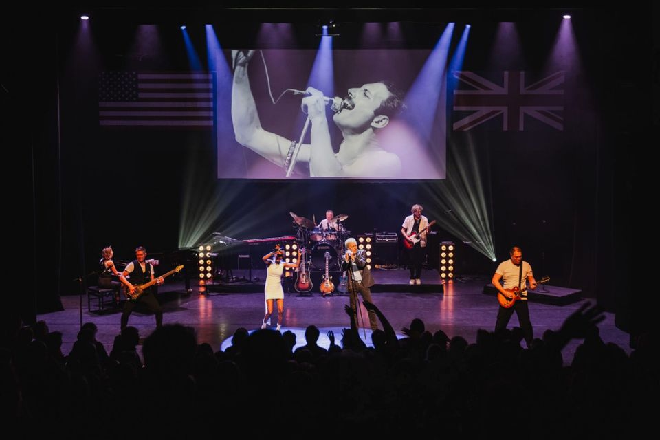 Foto van een podium met een 7-koppige band en instrumenten. Boven hen wordt Freddy Mercury getoond op een scherm.