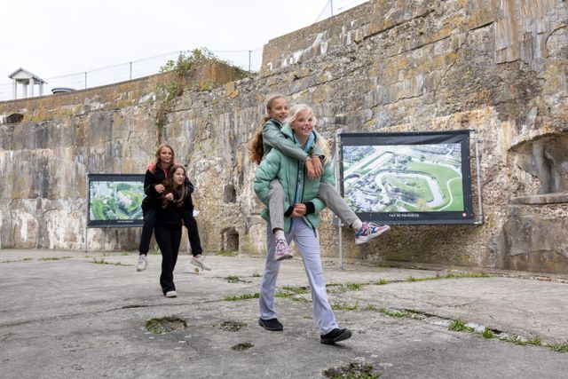 Vier kinderen lopen samen door de droge gracht rondom het fort op forteiland Pampus. Op de achtergrond zie je de robuuste wanden van het fort en staan grote foto's van een tentoonstelling. Twee kinderen dragen de andere twee kinderen op hun rug.