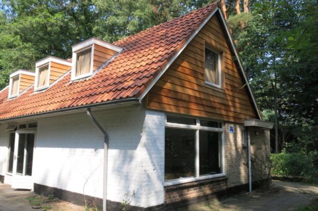 Vakantiehuis met een witte gevel, puntdak en drie dakkapellen in een bosrijke omgeving.