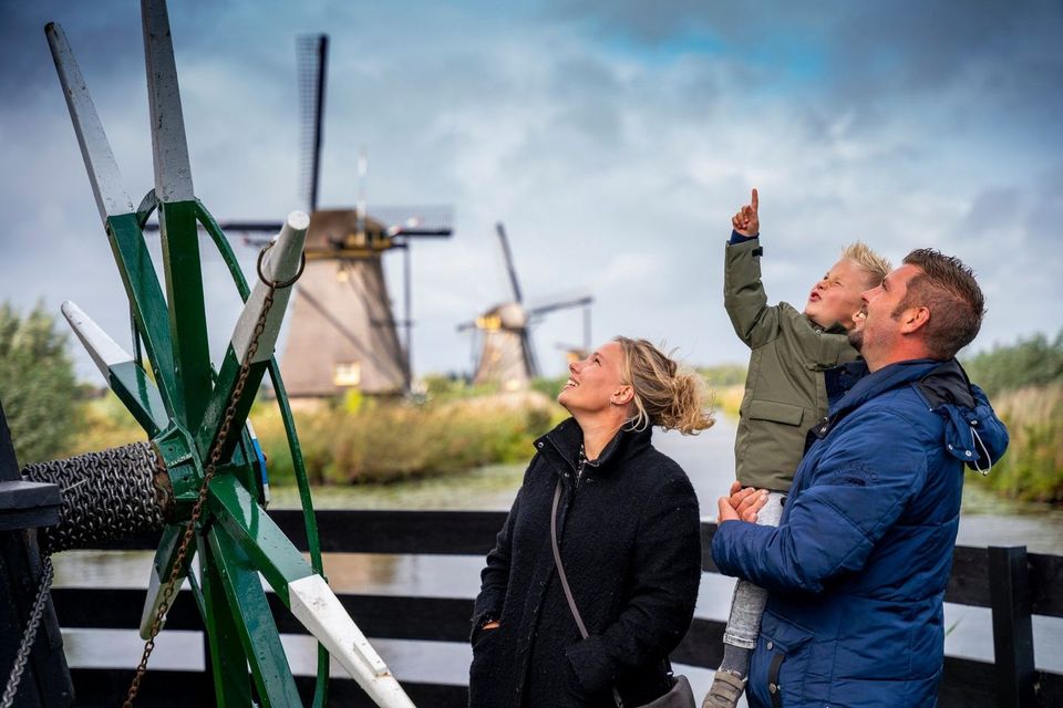Poldermolens in het Groene Hart, waar een gezin met een kind van dichtbij naar een molen kijkt met meerdere historische windmolens langs het water op de achtergrond.