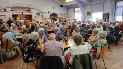 Grote groep senioren zit aan tafels in een buurthuiszaal en praat met elkaar tijdens een drukbezochte bijeenkomst met koffie en drankjes.