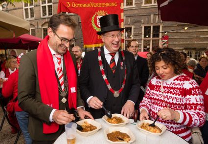 Burgermeester, voorzitter en een mevrouw eten hutspot aan een staan sta tafel.