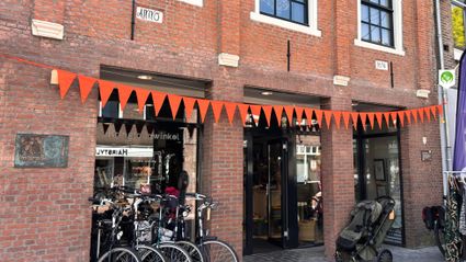 Voorgevel van kringloopwinkel De Terugwinning in het centrum van Woerden, met oranje vlaggetjes, fietsen voor de deur en kledingrekken buiten op straat.