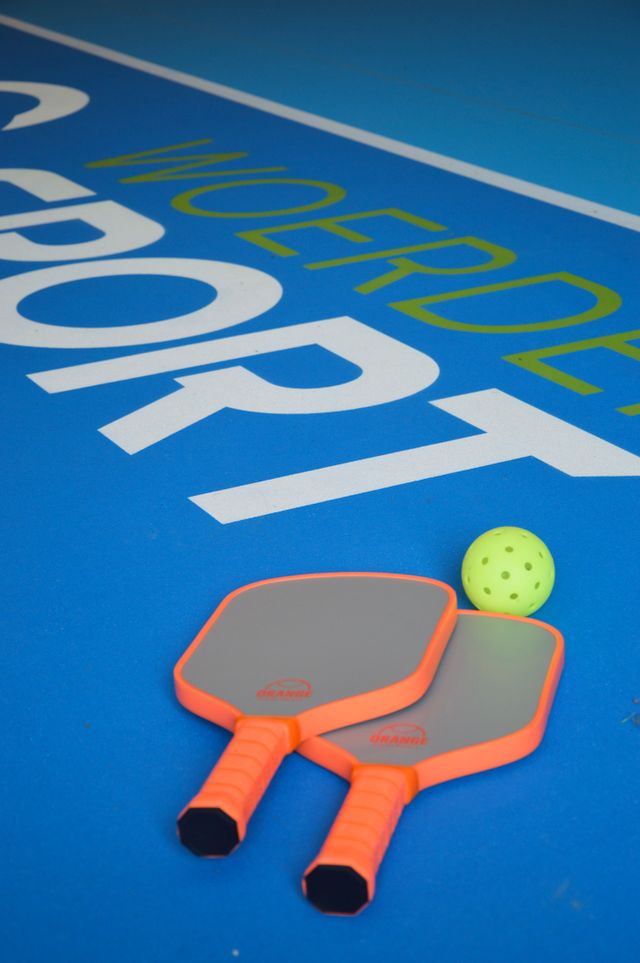 Pickleballbaan bij Zwembad H2O in Harmelen, Groene Hart, met twee pickleballpaddles en een gele geperforeerde bal op de blauwe sportvloer.