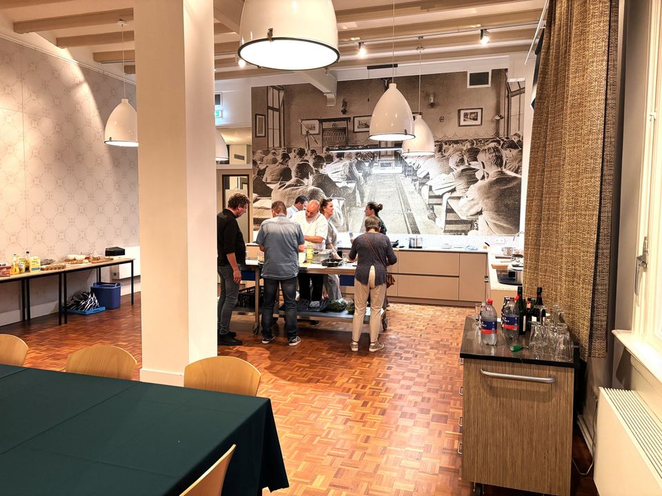 Vijf personen staan rond een rvs-werktafel in een open keuken en bereiden eten. Op de achtergrond het lange keukenblok en de grote zwart-wit muurfoto; rechts staan glazen en flessen klaar op een kar.