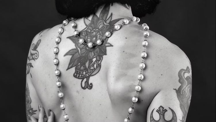 Zwart-wit foto van een vrouwenrug met tatoeages en een lange parelketting.