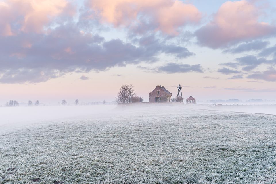 Een bevroren landschap onder een roze lucht. In de verte zie je een klein huisje op Schokland in de Noordoostpolder.