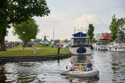 Een rubberboot met twee mannen en een jongetje vaart in de volle haven van Joure met op de achtergrond het zicht op een molen.