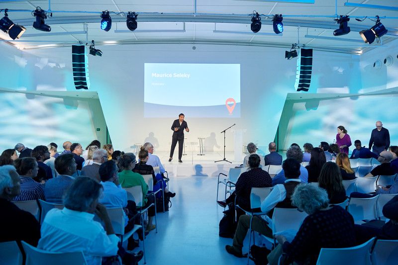 Een man geeft een presentatie met een beeldscherm achter zich. Links en rechts zit publiek.