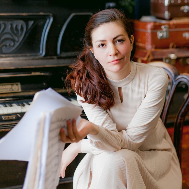 Foto van Ekaterina in het wit gekleed, zittend voor een piano en opgestapelde leren koffers.