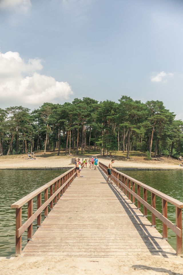 Een houten vlonder loopt over het water naar het zandstrand van het Henschotermeer bij Woudenberg. Op de achtergrond liggen recreanten tussen de hoge naaldbomen.