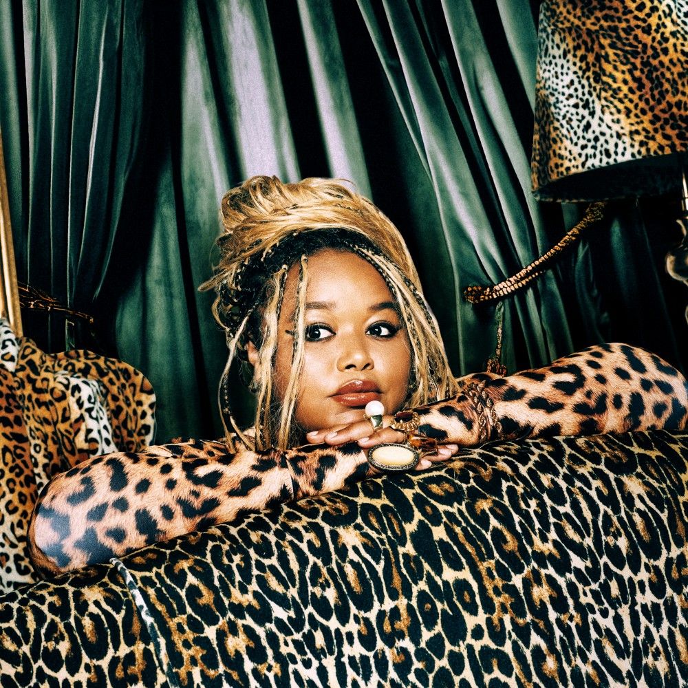 Foto van Sarah gehuld in luipaardprint kleding, tussen luipaardprint kleden en lamp, voor een donkergroen gordijn.