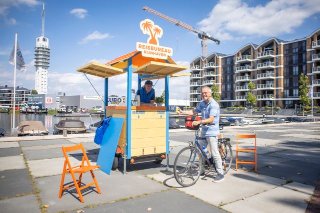 Een vrouw achter het reisbureau kraampje van Rijnhaven met een meneer op zijn fiets die daarnaast staat.