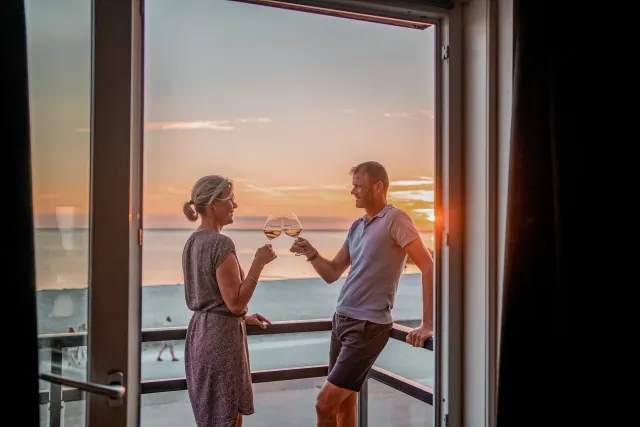 Een koppel proostend met een glas wijn op het balkon met uitzicht over het strand bij Strandhotel Vigilante in Makkum.