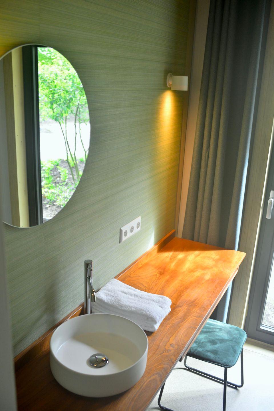 Detailfoto van een wastafel op een houten plank met een grote ronde spiegel erboven en blauw krukje.