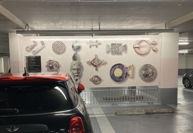 Bij een geparkeerde auto hangt een paneel met vergrote afbeeldingen van diverse vondsten: een olielampje, fibula's, metalen plaatjes.