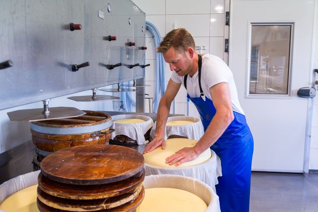 Twee vrouwelijke medewerkers staan lachend achter de toonbank kaas te snijden, in de kaaswinkel van Kaasboerderij Captein.