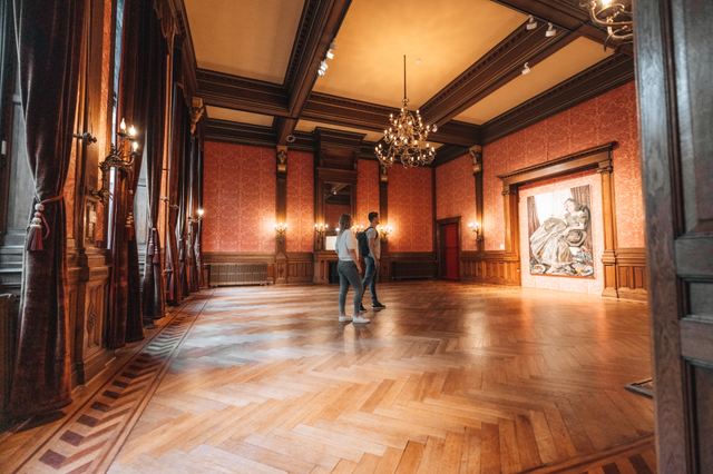 Een man en een vrouw staan in een zal verder lege zaal, met een prachtige houten vloer, naar een groot kunstwerk aan de muur te kijken.
