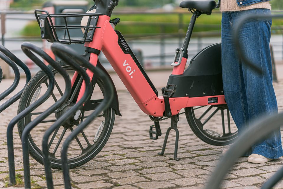 VOI deelfiets staat in een fietsenrek. Daarnaast zie je de benen van een vrouw in spijkerbroek.