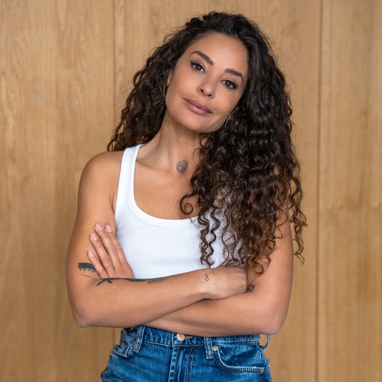 Foto van Fajah met lange donkere krullen, in een wit hemdje en spijkerbroek voor een houten paneel achtergrond.