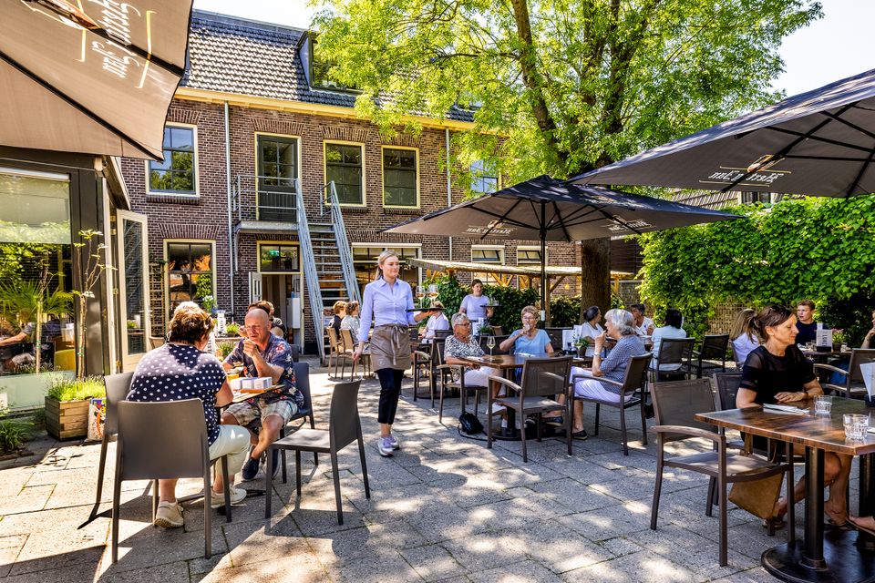 Zonnig terras van Hotel en Grand Café Broeck in Oudewater met gasten aan tafels onder parasols, bediend door personeel, gelegen in een groene binnentuin bij een historisch pand in het centrum van Oudewater.