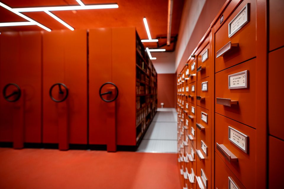 Oranje archiefkasten met gelabelde laden in een archiefdepot, naast verrijdbare stellingen, ingericht voor het systematisch opslaan en terugvinden van documenten.