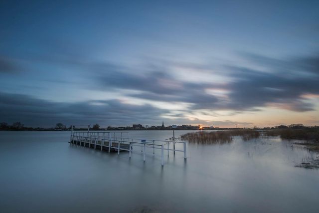 ChatGPT zei:

Natuurgebied Salmsteke in Lopik bij zonsondergang, met een witte steiger in het hoge water van de Lek, riet en overstroomde uiterwaarden, en in de verte de dorpssilhouetten met kerktoren onder dramatische wolken.