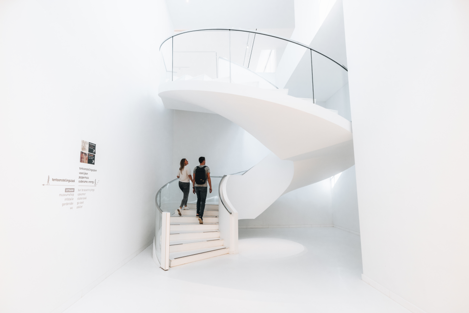 Een man en een vrouw beklimmen de witte wenteltrap in het Drents Museum in een ruimte met witte muren, een witte vloer en een wit plafond.