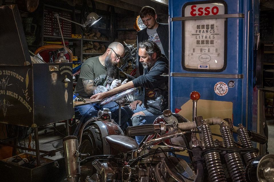 Werkplaatsfotografie tatoeage en motoren, mannen bezig met tatoeëren tussen motorfietsen in werkplaats, Woerden, Groene Hart, rauwe binnenruimte met industriële sfeer en focus op ambacht en detail.