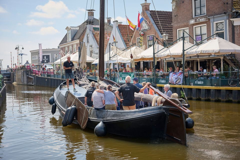Er vaart een authentieke zeilboot door de gracht van Lemmer. Er staat een man op de punt en er staan en zitten mensen in de boot. Op de achtergrond staan grachtenpandjes en terrassen met open parasols.