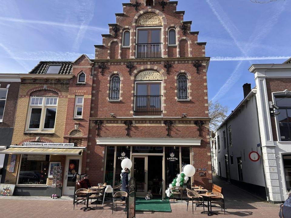 Historisch horecapand Barista Café Woerden in Woerden, Groene Hart, met zonnig terras, gasten aan tafels en feestelijke ballonnen bij de entree van het karakteristieke bakstenen pand.