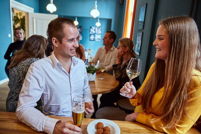 Twee mensen lachend aan een tafel met een glas bier, wijn en bitterballen bij Proeflokaal Het Gerechtshof in Sneek.