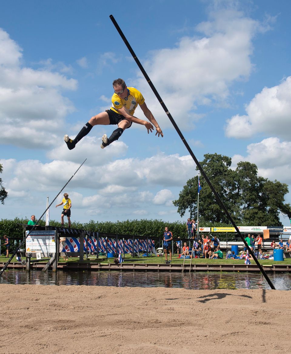 Man springt met een polsstok over het water tijdens het NK Polsstokverspringen, met toeschouwers op de achtergrond in Linschoten.