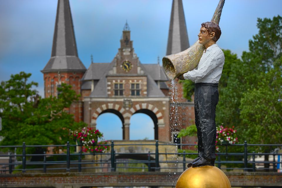Beeld van een man met een grote hoorn waar water uit kabbelt met op de achtergrond de Waterpoort van Sneek
