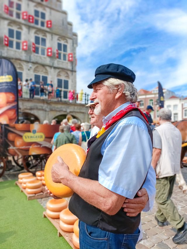 Man in traditionele kleding houdt een grote ronde kaas vast tijdens de kaasmarkt in Gouda, met historische gebouwen en stapels kazen op de achtergrond in het Groene Hart.