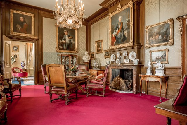 De rijk ingerichte rookkamer van Huis Doorn toont historische meubels, schilderijen en porselein. De houten lambrisering en rode vloer geven de ruimte een klassieke uitstraling.