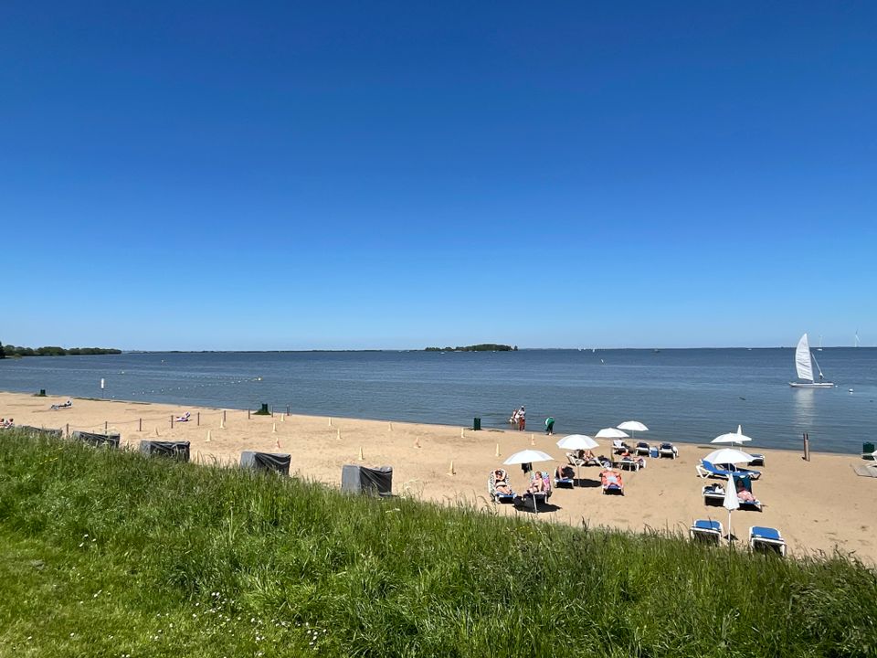Strand Muiderberg vanaf het land gezien, met op de achtergrond het IJmeer en een zeilbootje.