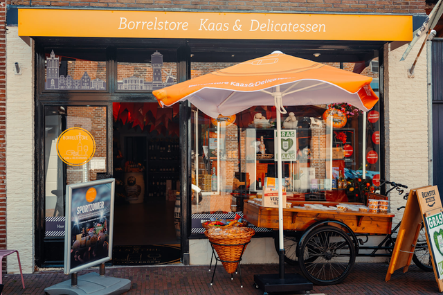 De voorzijde van een delicatessewinkel met voor de etalage een fietskar en parasol.