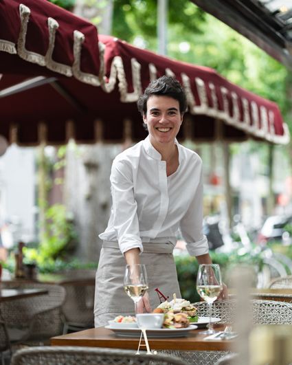 Serveerster in witte kleding op terras van restaurant AMBIANS. in Oisterwijk onder bordeauxrode parasol