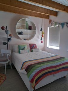 Sfeervolle slaapkamer met tweepersoonsbed, kleurrijke gehaakte sprei, sierkussens en ronde nis boven het hoofdbord, verlicht door een raam met rolgordijn.