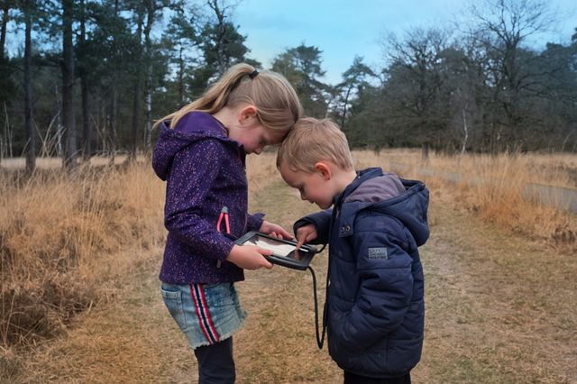 Der Bruder und die Schwester stehen in der Natur und schauen auf ein Tablet, wohin sie gehen sollen