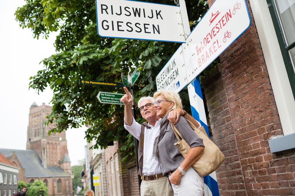 Een man en vrouw staan onder een wegwijzer in een oude binnenstad. De man heeft zijn arm om de vrouw heen en wijst met zijn hand iets aan, waar de vrouw naar kijkt.