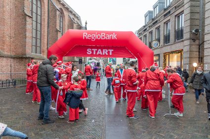 Een vrolijk en energiek tafereel in de Woerdense binnenstad, waar deelnemers in rood-witte kerstoutfits zich verzamelen onder de grote startboog van de Santa Run. Tussen de historische gebouwen hangt een uitbundige sfeer van plezier, saamhorigheid en voorpret, terwijl jong en oud zich klaarmaakt voor een feestelijke hardlooptocht door de stad.