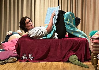 Foto van een vrouw, zij ligt op een rood bed met een dekentje en een boek. Onder het bed ligt een knuffelmonster en om haar heen nog meer knuffeldiertjes.