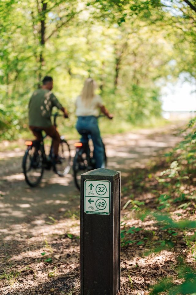 Ein Paar radelt durch den Wald. Im Vordergrund steht ein Wegweiser des Knotenpunktsystems.