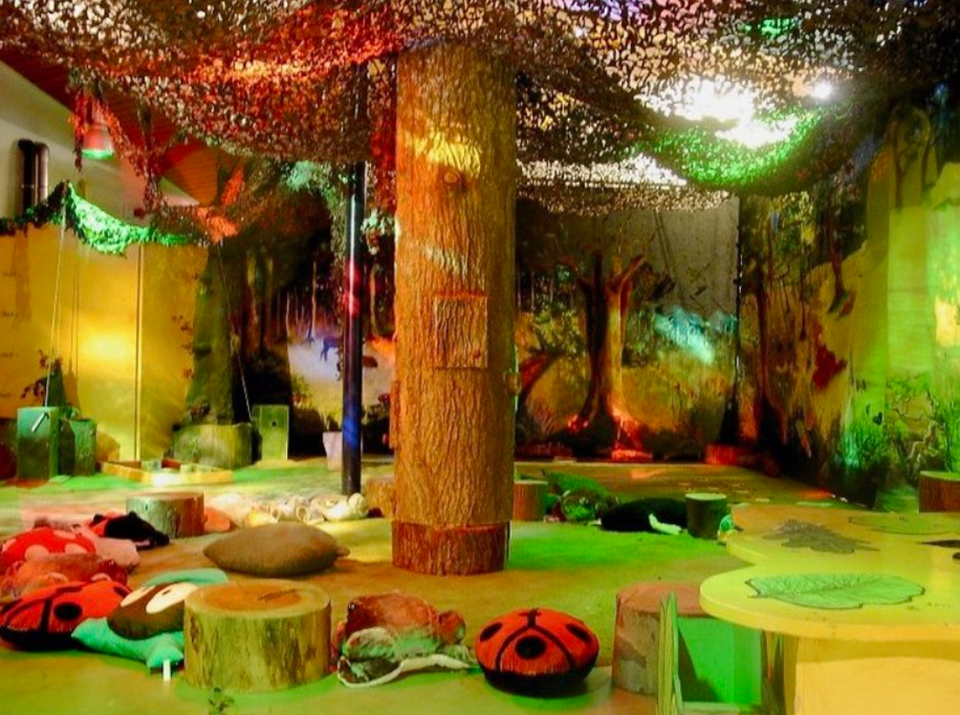 Sfeervol ingerichte binnenruimte in bos-thema met boomstammen, kussens en zachte zitplekken. Gekleurd licht schijnt door een net aan het plafond, met wandschilderingen van bomen en natuur voor een magische, rustige ambiance.