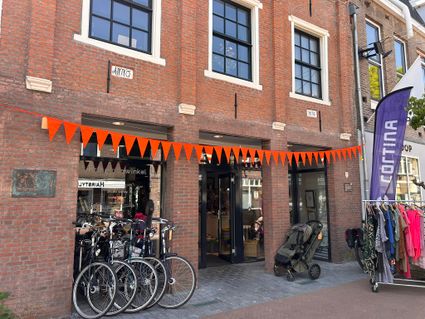 Voorgevel van kringloopwinkel De Terugwinning in het centrum van Woerden, met oranje vlaggetjes, fietsen voor de deur en kledingrekken buiten op straat.